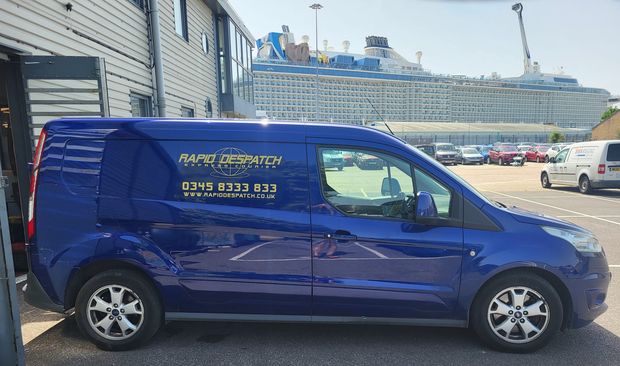 A blue Rapid Despatch van parked by a quayside, with a cruise ship behind
