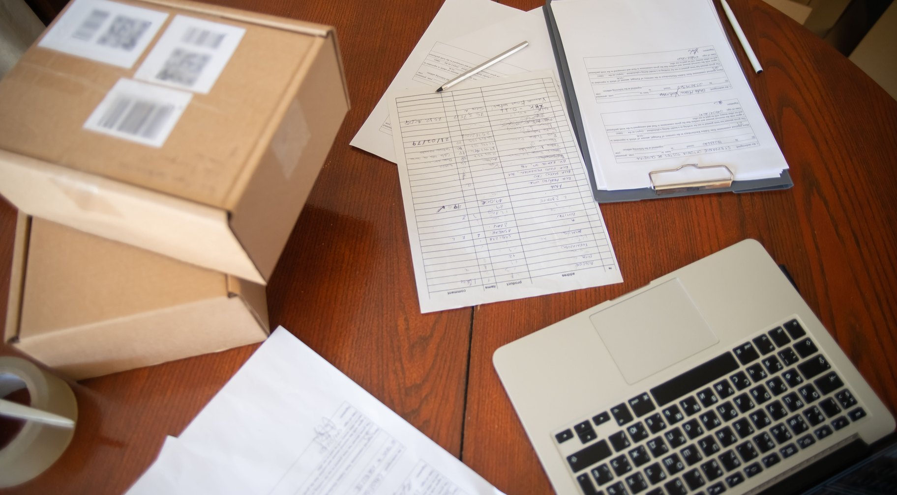 Shipping paperwork, packages and a laptop laid out on a desk